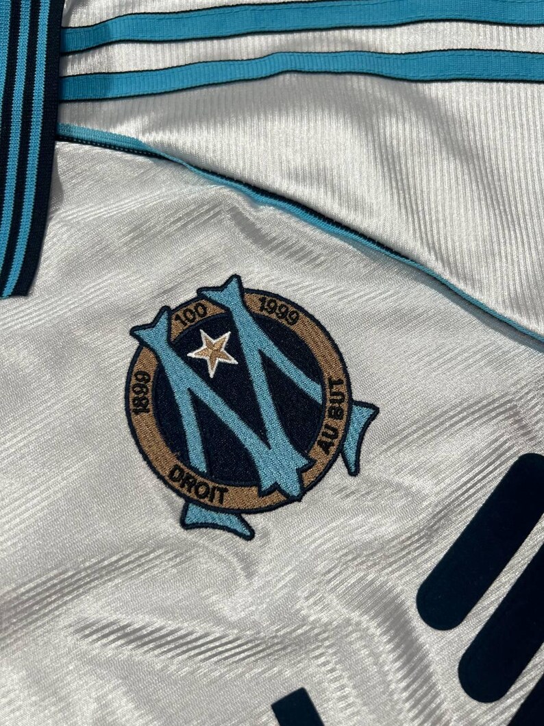 Peut inclure: Gros plan sur un maillot de football blanc et bleu clair. Le maillot pr&eacute;sente un logo d'&eacute;quipe avec le texte "1899", "1999" et "Droit". Le maillot a des accents bleu clair et bleu marine.