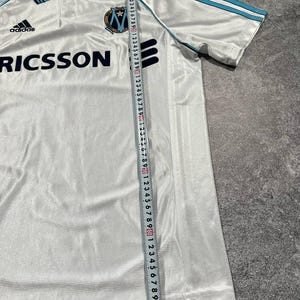 Peut inclure: Maillot de football Adidas blanc avec des d&eacute;tails bleu clair sur le col et les manches. Il pr&eacute;sente le logo Adidas, le mot "RICSSON" et un &eacute;cusson d'&eacute;quipe. Une r&egrave;gle est utilis&eacute;e pour mesurer le maillot.