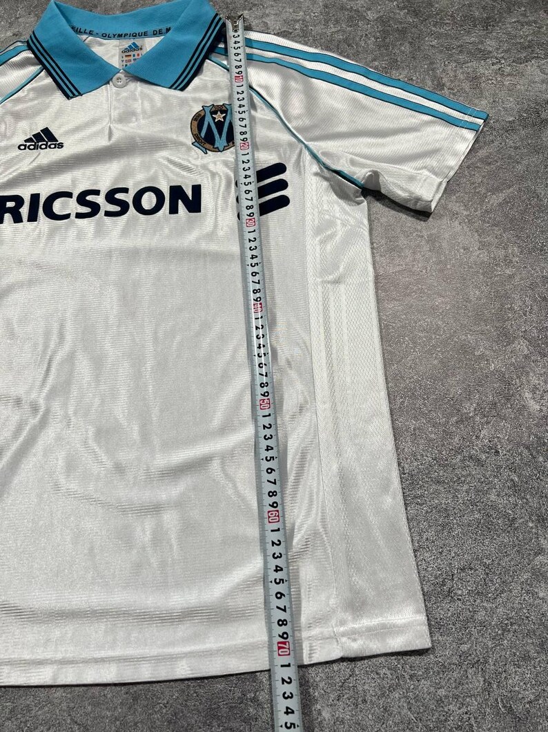Peut inclure: Maillot de football Adidas blanc avec des d&eacute;tails bleu clair sur le col et les manches. Il pr&eacute;sente le logo Adidas, le mot "RICSSON" et un &eacute;cusson d'&eacute;quipe. Une r&egrave;gle est utilis&eacute;e pour mesurer le maillot.