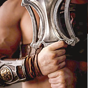 May include: A silver-colored sword with a detailed handle is held by a person. The sword has a unique design with curved elements. The person is wearing a brown leather arm guard with gold accents. The image is set against a dark background.
