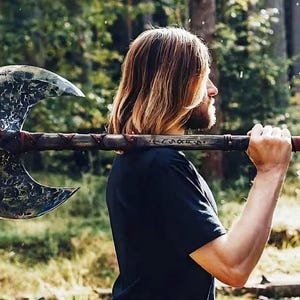 May include: A large, double-bladed axe with a weathered, textured blade and a wooden handle. The handle is wrapped with red leather near the grip. The axe is held over the shoulder against a blurred forest background.