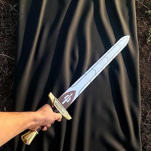May include: A toy sword with a white blade and a gold hilt. The hilt has a brown grip and a decorative emblem. The sword is held against a black background.