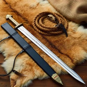 May include: A Viking-style sword with a silver blade and a gold-colored hilt, resting in a black leather sheath. The blade has etched inscriptions. The sword is displayed on a brown fur rug, with a coiled rope and a small skull in the background.