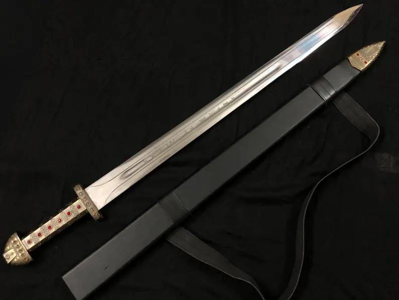 May include: A silver sword with a decorative gold and red handle and a black sheath. The sword's blade is long and sharp, and the sheath has a strap for carrying. The sword is likely a replica or decorative item.
