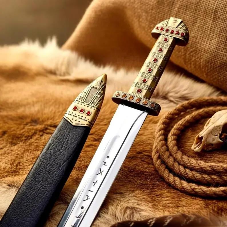 May include: A decorative sword with a silver blade and a gold-colored handle adorned with red gemstones. The sword is displayed with its black leather sheath on a textured brown surface, alongside a coiled rope and a skull.