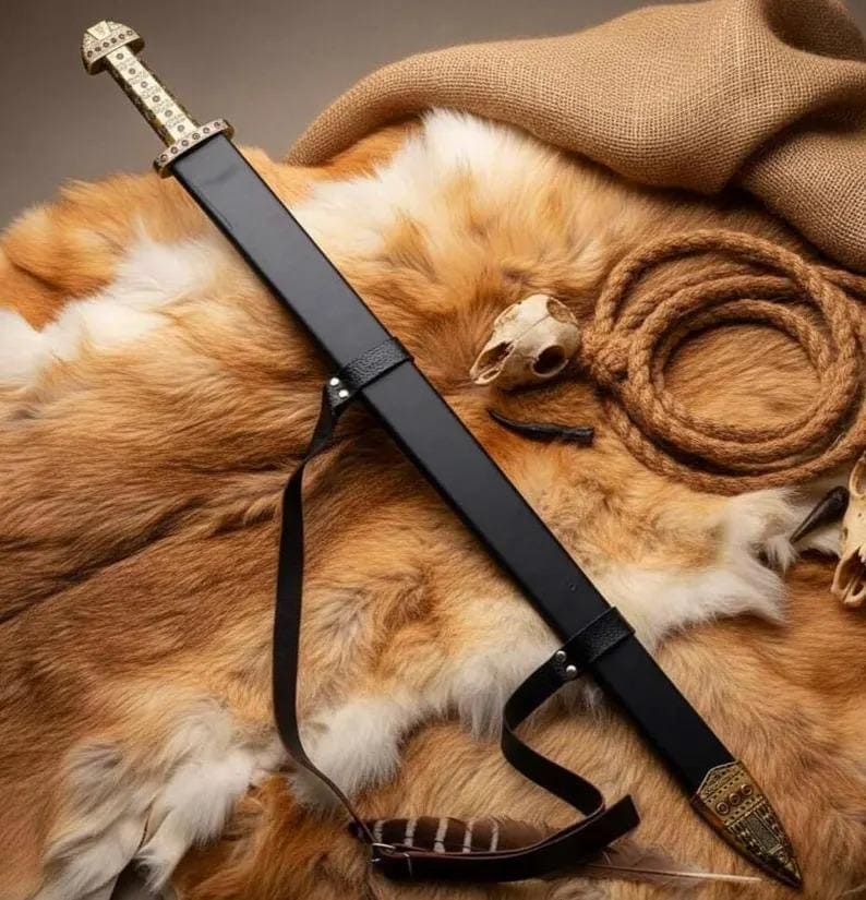 May include: A black sword with a gold-colored hilt and scabbard tip rests on a brown fur surface. The sword has a black leather strap and is accompanied by a coiled rope and animal skull, suggesting a historical or fantasy theme.