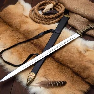May include: A Viking-style sword with a silver blade and a decorative gold handle, resting in a black sheath. The sword is displayed on a brown fur pelt, with a leather strap, rope, and a feather.