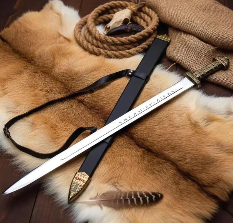 May include: A Viking-style sword with a silver blade and a decorative gold handle, resting in a black sheath. The sword is displayed on a brown fur pelt, with a leather strap, rope, and a feather.