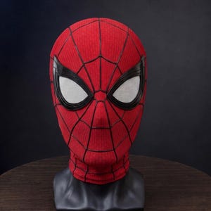 May include: A red fabric mask featuring a black web pattern across the surface. The mask includes large, angular eye lenses with white mesh inserts framed by thick black borders, designed to cover the entire head and neck area completely.
