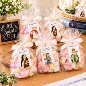 Op de afbeelding: Vijf doorzichtige plastic traktatiezakjes gevuld met pastelkleurige marshmallows en macarons, elk gestrikt met een crèmekleurig lint en een label met een heiligenillustratie en teksten zoals Be holy, Be kind, Be a Saint en Shine for Jesus.