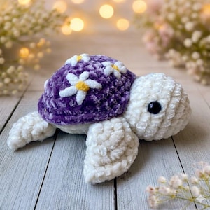 Peut inclure: Une tortue crochetée à la main. La carapace violette est ornée de marguerites blanches et jaunes. La tête, les nageoires et le dessous sont blancs. Posée sur une surface en bois de couleur claire.