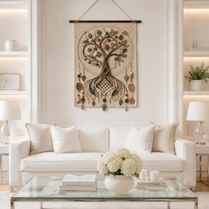 May include: A rectangular cream-colored crochet wall hanging featuring a central tree design with intricate brown and green textured branches, leaves, and roots. The piece is suspended from a dark wooden rod and includes dangling beaded accents.