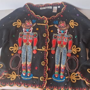 以下が含まれることがあります： 前面に2体のカウボーイのくるみ割り人形が刺繍された、黒の長袖カーディガン。セーターはオレンジとグリーンのトリムが付いたスカラップエッジで、ゴールドトーンのボタン、ゴールドのロープのアクセント、星、スパンコールで装飾されています。