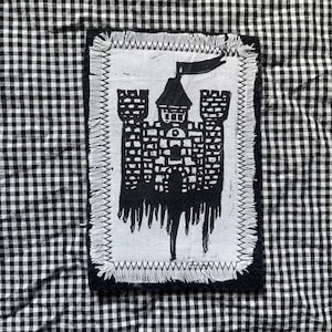 May include: A black and white patch featuring a castle design with a flag. The patch is rectangular with a white background and a black border. The castle is black and has a detailed design. The patch is sewn onto a black and white checkered fabric.