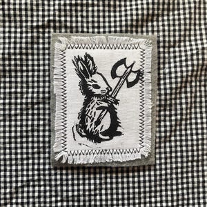 May include: A black and white patch featuring a rabbit holding an axe. The rabbit is in the center of the patch, with a gray border and white stitching. The background is a black and white checkered pattern.