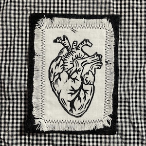 May include: A rectangular patch with a black and white anatomical heart design. The heart is printed in black ink on a white fabric, framed by a black border and a white stitched edge. The background is a black and white checkered pattern.