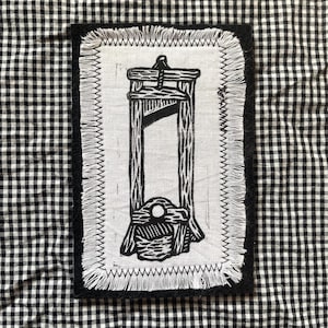 May include: A rectangular patch with a black border and a white center featuring a black illustration of a guillotine. The patch has a frayed edge and is set against a black and white checkered background.