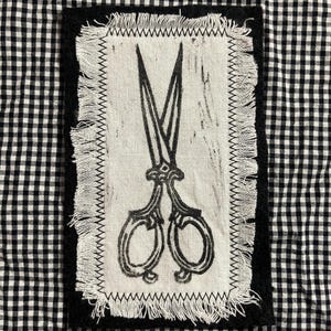 May include: A black and white fabric patch featuring a detailed illustration of scissors. The scissors are printed in black on a white background, with a frayed edge. The patch is set against a black and white checkered fabric.