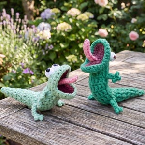 Puede incluir: Dos juguetes de lagarto verde tejidos a ganchillo con bocas abiertas y lenguas rosas. Los lagartos tienen ojos blancos y están colocados sobre una superficie de madera. El fondo presenta un jardín con flores y vegetación.