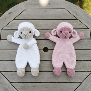 Puede incluir: Dos mu&ntilde;ecos de cordero hechos a mano con ganchillo. Uno es blanco con detalles beige, el otro es rosa con detalles rosa m&aacute;s oscuros. Ambos tienen los brazos extendidos y est&aacute;n sobre una superficie de madera. Los mu&ntilde;ecos est&aacute;n dise&ntilde;ados para parecer ovejas.