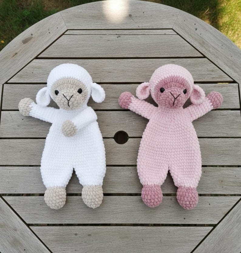 Puede incluir: Dos mu&ntilde;ecos de cordero hechos a mano con ganchillo. Uno es blanco con detalles beige, el otro es rosa con detalles rosa m&aacute;s oscuros. Ambos tienen los brazos extendidos y est&aacute;n sobre una superficie de madera. Los mu&ntilde;ecos est&aacute;n dise&ntilde;ados para parecer ovejas.