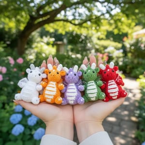 May include: A collection of five colorful crochet dragon keychains. The dragons are white, orange, purple, green, and red, each with unique wing and belly patterns. They are held in open hands against a blurred garden backdrop.