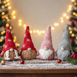 Pode incluir: Quatro gnomos de crochê feitos à mão, com chapéus pontudos longos e barbas brancas. Os gnomos são nas cores vermelho, rosa e cinza, com detalhes de flocos de neve brancos em seus chapéus. Eles estão dispostos em uma superfície de madeira.