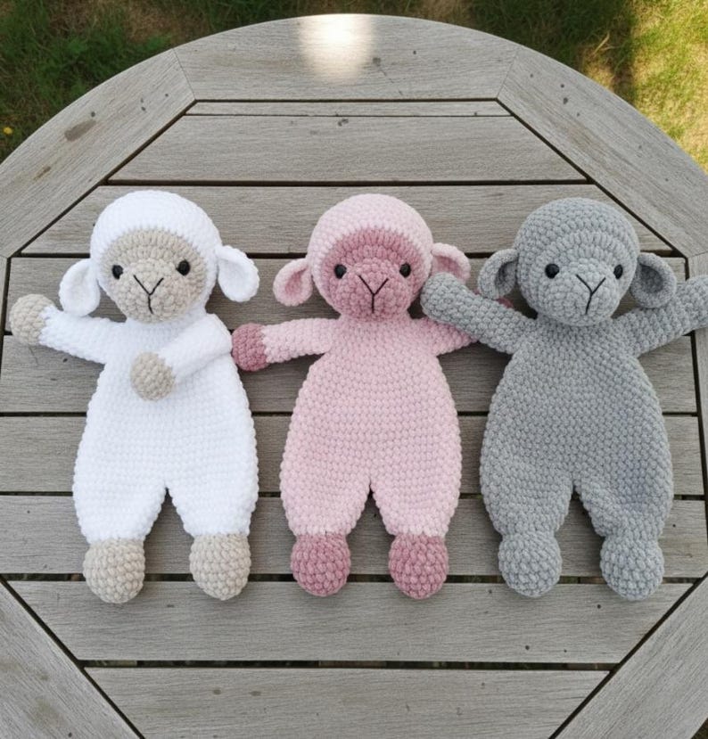 Puede incluir: Tres mu&ntilde;ecos de cordero hechos a mano con ganchillo en blanco, rosa y gris. Cada cordero tiene una cara detallada, orejas peque&ntilde;as y brazos extendidos. Los mu&ntilde;ecos est&aacute;n colocados sobre una superficie de madera, mostrando su aspecto suave y texturizado.