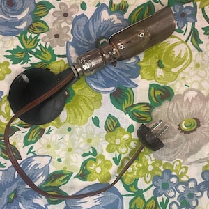 May include: A vintage, small, black lamp with a silver-coloured bulb housing and a brown cord. The lamp has a round base and a plug at the end of the cord. The lamp is on a floral patterned fabric.