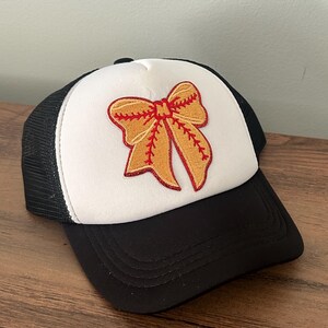 May include: Black and white trucker hat with a white front panel featuring an embroidered softball bow in shades of gold and red. The hat has a curved brim and a mesh back.