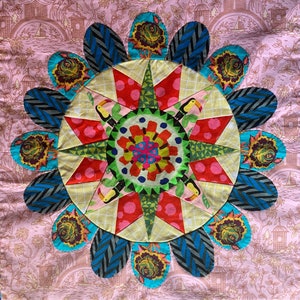 May include: A colorful patchwork quilt block with a starburst design in the center. The block is made up of various fabrics with floral patterns and geometric shapes. The background is a light pink.