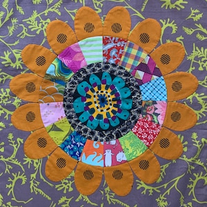 May include: A colorful patchwork quilt block in the shape of a sunflower with a central circular design. The sunflower is made of various fabrics in shades of red, blue, green, yellow, and orange.