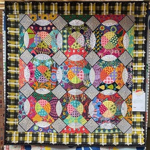 Könnte beinhalten: Ein farbenfroher Quilt mit einem Patchwork-Design. Der Quilt zeigt ein sich wiederholendes Muster aus Kreisen, die mit verschiedenen gemusterten Stoffen in leuchtenden Farben gefüllt sind. Der Rand ist schwarz-gelb kariert.