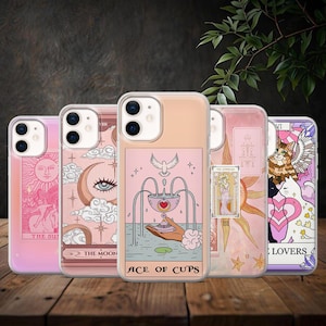 May include: Five phone cases with tarot card illustrations in pink, peach, and purple. The designs include The Sun, The Moon, Ace of Cups, The Empress, and The Lovers tarot cards. The cases are displayed on a wooden surface.