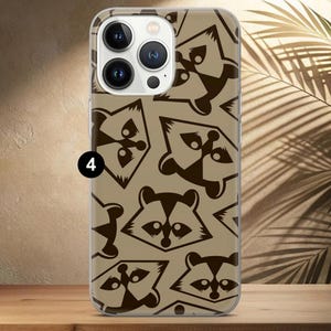 May include: A clear phone case featuring a repeating pattern of brown raccoon faces and abstract shapes on a tan background. The case has a clear border and covers the back and sides of the phone.