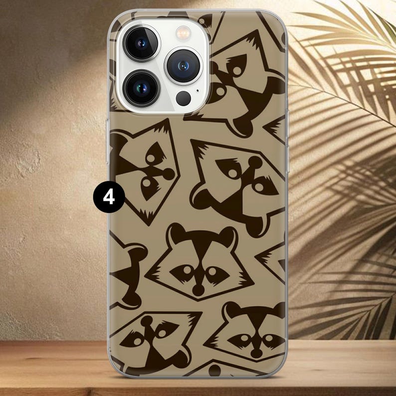 May include: A clear phone case featuring a repeating pattern of brown raccoon faces and abstract shapes on a tan background. The case has a clear border and covers the back and sides of the phone.