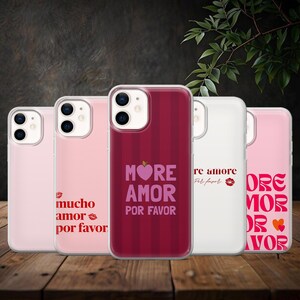 Op de afbeelding: Een verzameling van vijf transparante telefoonhoesjes met verschillende ontwerpen. De hoesjes tonen zinnen als "More Amor Por Favor" en "Mucho Amor Por Favor" in roze en rode lettertypen. De hoesjes worden gepresenteerd op een houten oppervlak.