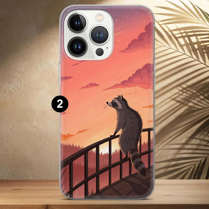 May include: A phone case featuring a raccoon on a bridge, set against a sunset sky. The raccoon is gray and white, with a striped tail. The background includes a forest and a vibrant orange and pink sky.