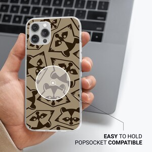 May include: A clear phone case with a repeating pattern of brown raccoon faces on a tan background. The case is held in a hand and is labeled "EASY TO HOLD POPSOCKET COMPATIBLE".