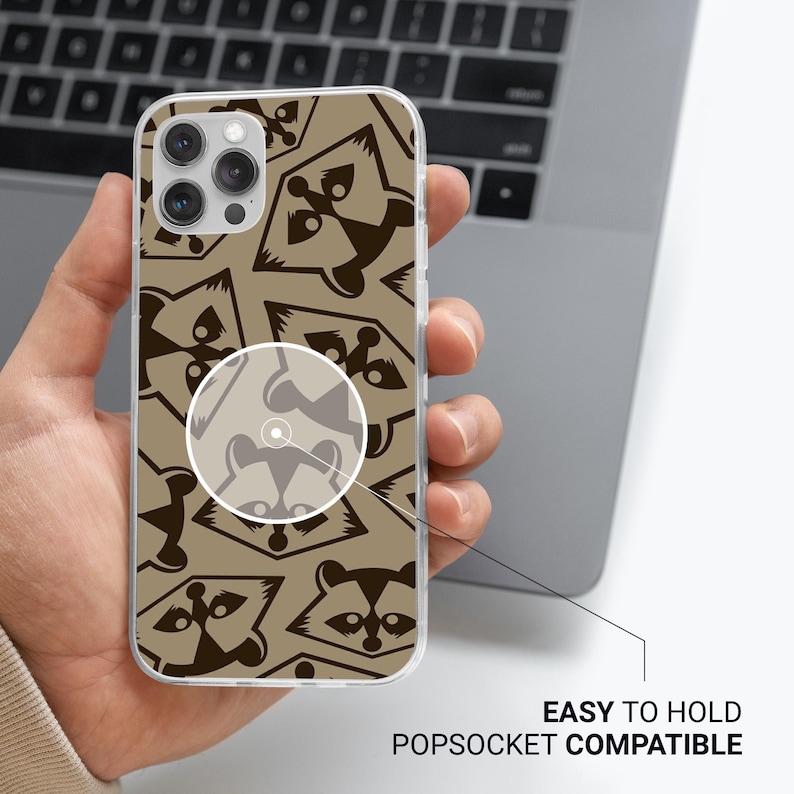 May include: A clear phone case with a repeating pattern of brown raccoon faces on a tan background. The case is held in a hand and is labeled "EASY TO HOLD POPSOCKET COMPATIBLE".