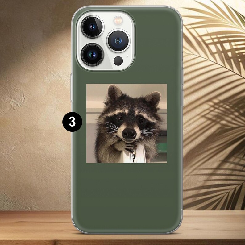 May include: Green phone case featuring a square image of a raccoon holding a white object. The raccoon has black and gray fur with a white face and is looking directly at the viewer. The phone case has a clear border and a camera module.