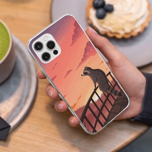 May include: Clear phone case featuring a raccoon illustration. The design shows a raccoon on a fence, with a sunset backdrop in shades of orange, pink, and purple. The case is held in a hand, with a blurred background.