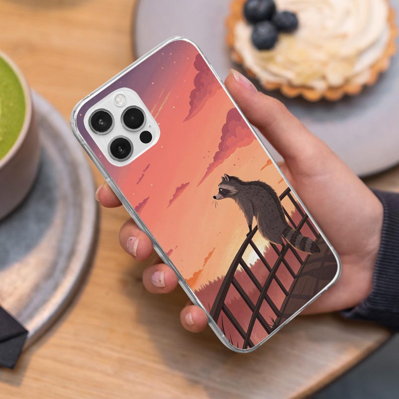 May include: Clear phone case featuring a raccoon illustration. The design shows a raccoon on a fence, with a sunset backdrop in shades of orange, pink, and purple. The case is held in a hand, with a blurred background.