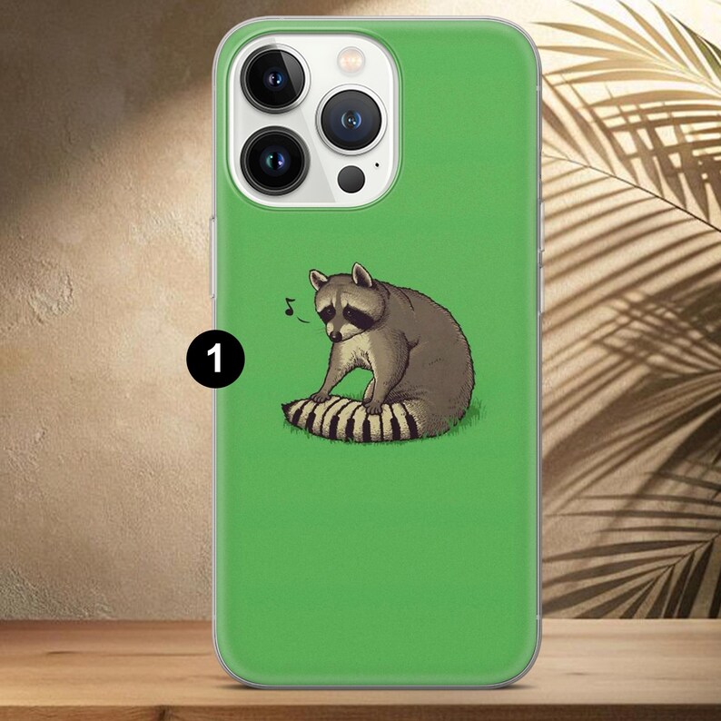 May include: Green phone case featuring a raccoon illustration. The raccoon is depicted in shades of gray and brown, sitting on a striped tail. A musical note floats above the raccoon. The case has a clear border and a camera cutout.