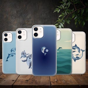 Op de afbeelding: Een verzameling van vijf telefoonhoesjes met dolfijnontwerpen. De hoesjes hebben verschillende tinten blauw, wit en beige, met illustraties van zwemmende en springende dolfijnen. De hoesjes worden op een houten oppervlak gepresenteerd.