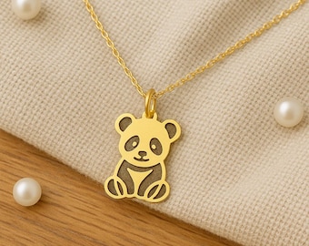 May include: A gold-colored panda pendant necklace. The pendant features a panda bear design with black accents. The necklace is displayed on a beige fabric with three pearls and a wooden surface.