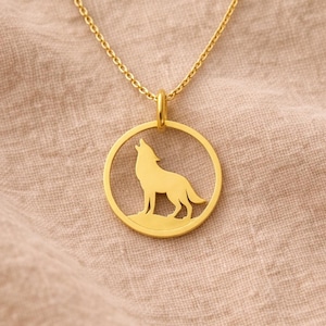 Può includere: Collana color oro con la silhouette di un lupo che ulula all'interno di un ciondolo circolare. Il ciondolo è sospeso a una delicata catena. Il lupo è raffigurato in piedi su una piccola piattaforma. Lo sfondo è un tessuto neutro e strutturato.