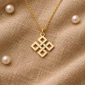 Puede incluir: Un collar de tono dorado con un colgante de diseño geométrico en forma de nudo infinito cuadrado. El colgante cuelga de una delicada cadena dorada y la pieza está rodeada por varias perlas blancas pequeñas y redondas.