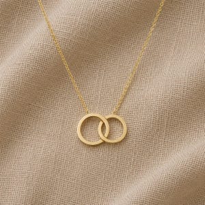 Può includere: Una collana color oro con un ciondolo composto da due anelli circolari intrecciati. Il ciondolo è sospeso a una sottile e delicata catenina color oro. Il design è costituito da due cerchi lisci e lucidi collegati insieme al centro.