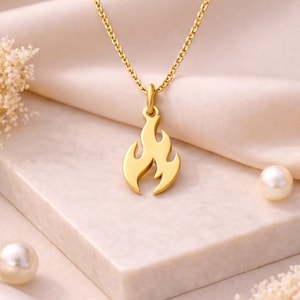 May include: A gold necklace featuring a flame-shaped pendant. The pendant is a stylized flame design, and the chain is a delicate link style. The necklace is presented on a light-coloured surface with pearls and floral accents.